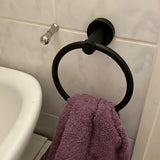 Une serviette violette est suspendue à l'anneau porte-serviettes Elarbo Black de yessential.fr, monté sur un mur carrelé à côté d'un lavabo et d'un crochet argenté, ajoutant une touche peu encombrante à la salle de bain.
