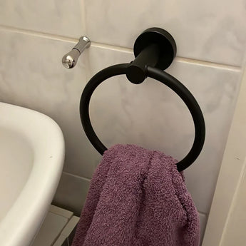 Une serviette violette est suspendue à l'anneau porte-serviettes Elarbo Black de yessential.fr, monté sur un mur carrelé à côté d'un lavabo et d'un crochet argenté, ajoutant une touche peu encombrante à la salle de bain.