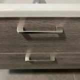 Un gros plan de deux poignées Ramiro Chrome Deluxe de yessential.fr sur un tiroir d'armoire de salle de bain à structure en bois, avec un rebord d'évier blanc au-dessus et des carreaux gris élégants au sol.