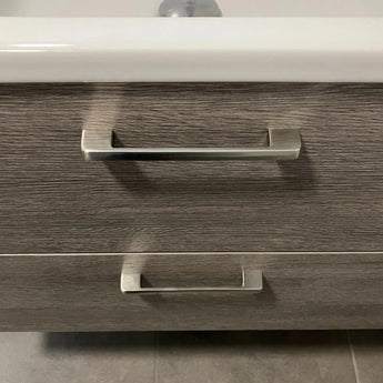 Un gros plan de deux poignées Ramiro Chrome Deluxe de yessential.fr sur un tiroir d'armoire de salle de bain à structure en bois, avec un rebord d'évier blanc au-dessus et des carreaux gris élégants au sol.