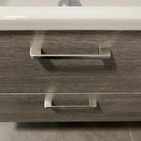 Un gros plan de deux poignées Ramiro Chrome Deluxe de yessential.fr sur un tiroir d'armoire de salle de bain à structure en bois, avec un rebord d'évier blanc au-dessus et des carreaux gris élégants au sol.