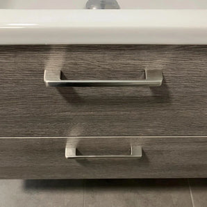 Un gros plan de deux poignées Ramiro Chrome Deluxe de yessential.fr sur un tiroir d'armoire de salle de bain à structure en bois, avec un rebord d'évier blanc au-dessus et des carreaux gris élégants au sol.