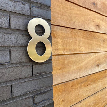 Un numéro 8 métallique Adamo de 15 cm en acier inoxydable de yessential.fr est monté sur un mur de briques sombres à côté d'un mur de panneaux de bois.
