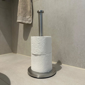 Un porte-rouleau de papier toilette autoportant yessential.fr Jurado en acier inoxydable se trouve sur le sol carrelé d'une salle de bain avec deux rouleaux de papier toilette blanc. Une serviette beige est suspendue au-dessus et à gauche de cet accessoire de toilette élégant.