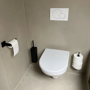 Une salle de bains moderne et minimaliste comprend des toilettes murales blanches avec de luxueux accessoires yessential.fr Vero Black Deluxe, une brosse de toilette noire, un porte-rouleau de papier toilette noir sur le mur de gauche et des rouleaux de rechange empilés sur un support carré à droite. Murs et sol gris clair.