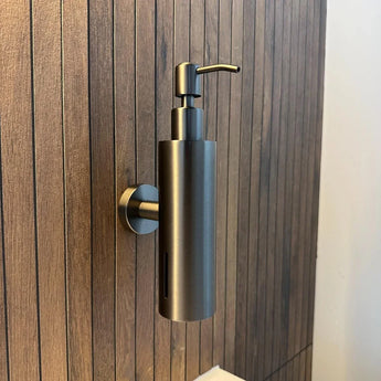 La pompe à savon Alberto Gunmetal de yessential.fr est un élégant distributeur de savon mural cylindrique en bronze à canon, monté sur un mur de salle de bain avec des panneaux verticaux en bois et équipé d'une pompe élégante sur le dessus pour une utilisation facile.