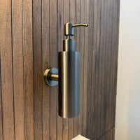La pompe à savon Alberto Gunmetal de yessential.fr est un élégant distributeur de savon mural cylindrique en bronze à canon, monté sur un mur de salle de bain avec des panneaux verticaux en bois et équipé d'une pompe élégante sur le dessus pour une utilisation facile.