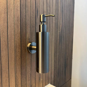 La pompe à savon Alberto Gunmetal de yessential.fr est un élégant distributeur de savon mural cylindrique en bronze à canon, monté sur un mur de salle de bain avec des panneaux verticaux en bois et équipé d'une pompe élégante sur le dessus pour une utilisation facile.