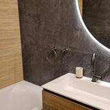Salle de bain moderne avec un miroir autour lumineux, un lavabo blanc, un robinet noir mat et l'anneau porte-serviettes Elarbo Gunmetal de yessential.fr comme accessoire. Les textures rugueuses de couleur beige et grise complètent le look ; baignoire à gauche.