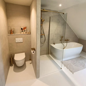 Salle de bain moderne avec murs beiges, toilettes flottantes, porte-rouleau de papier toilette Tazio Gold de yessential.fr, douche en verre, baignoire autoportante blanche, tapis clair, petits vases, brosse de toilette et œuvres d'art encadrées sur une étagère.