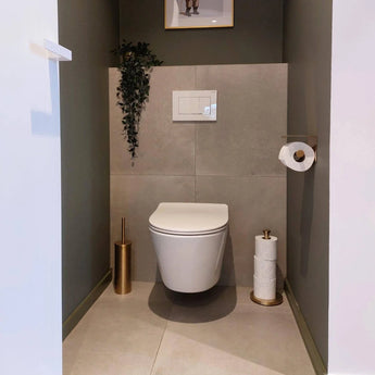 Une salle de bain moderne et minimaliste avec des toilettes murales, un porte-rouleau de papier toilette Tazio Gold de yessential.fr, une brosse et un support de toilette dorés assortis, du papier toilette mural, des rouleaux supplémentaires sur support et une plante suspendue pour la décoration.
