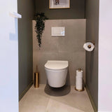 Une salle de bain moderne et minimaliste avec des toilettes suspendues, l'ensemble de brosses de toilettes Bruno Gold de yessential.fr, des rouleaux de rechange dans des supports dorés, une plante sur une étagère et du papier toilette sur un support doré.