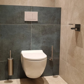 Une toilette murale moderne avec un bouton à double chasse au-dessus. Sur la droite se trouve un élégant porte-rouleau de papier toilette Tazio en acier inoxydable de yessential.fr, tandis que sur la gauche se trouve un porte-brosse de toilette, le tout contre des murs et un sol carrelés gris.