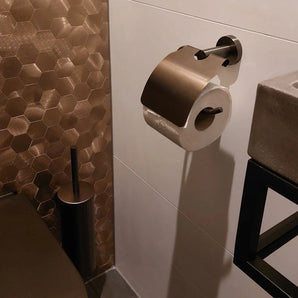 Une salle de bain moderne avec des carreaux hexagonaux en bronze, l'ensemble de toilettes Fabiano Gunmetal de yessential.fr, un porte-rouleau de papier toilette en métal avec rouleau, des accessoires assortis, ainsi qu'un lavabo gris et un porte-serviettes noir sur la photo.