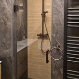 Une douche moderne avec les luminaires Elarbo Gunmetal de yessential.fr, une porte dans le mur, un mur vertical beige texturé, les carreaux avec la pierre gris foncé, les accessoires pour la salle de bain avec une porte-serviettes ronde supplémentaire et un radiateur avec serviettes à nez blanc.