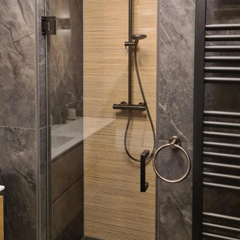 Une douche moderne avec les luminaires Elarbo Gunmetal de yessential.fr, une porte dans le mur, un mur vertical beige texturé, les carreaux avec la pierre gris foncé, les accessoires pour la salle de bain avec une porte-serviettes ronde supplémentaire et un radiateur avec serviettes à nez blanc.