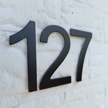 Numéro de maison Animo Black 7,6 cm (sans perceuse) de yessential.fr en noir sur un mur de briques blanches.