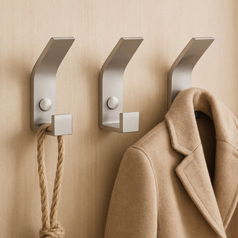 Trois crochets muraux Huso en acier inoxydable de yessential.fr, au design minimaliste et élégant, maintiennent un manteau beige et une corde épaisse contre un mur marron clair.