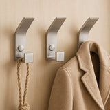 Trois crochets yessential.fr 10x Huso en acier inoxydable sont montés sur un mur beige : une corde épaisse pend au crochet de gauche, un manteau marron au crochet de droite et le crochet du milieu est vide.