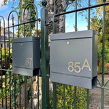 Deux boîtes aux lettres gris foncé avec les numéros 87 et 85A yessential.fr Animo en acier inoxydable 7,6 cm (sans perçage) sont montées à l'extérieur sur une clôture métallique noire, avec des arbres et des maisons en arrière-plan.