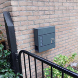 La boîte aux lettres Nevio Anthracite de yessential.fr portant le numéro 71 est montée sur un mur de briques, à côté de plantes vertes et d'une clôture en métal noir.