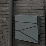 La boîte aux lettres Sergio Anthracite de yessential.fr, aux lignes épurées, est montée sur un mur de briques sombres à côté d'un tuyau d'évacuation gris assorti.
