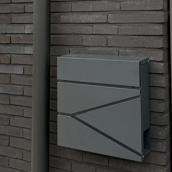La boîte aux lettres Sergio Anthracite de yessential.fr, aux lignes épurées, est montée sur un mur de briques sombres à côté d'un tuyau d'évacuation gris assorti.
