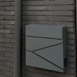 La boîte aux lettres Sergio Anthracite de yessential.fr, aux lignes épurées, est montée sur un mur de briques sombres à côté d'un tuyau d'évacuation gris assorti.