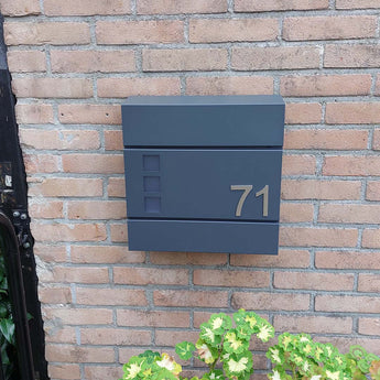 Le numéro Animo en acier inoxydable de 7,6 cm (sans perçage) de yessential.fr apparaît sous la forme « 71 » sur une boîte aux lettres moderne gris foncé, montée sur un mur de briques au-dessus de plantes vertes et jaunes.