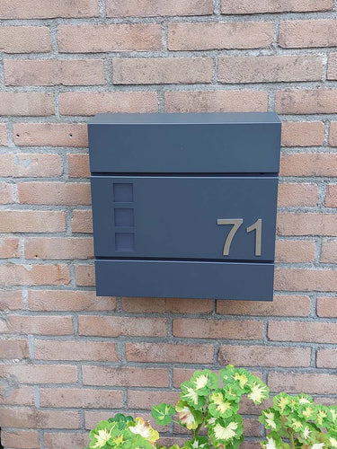 Le numéro Animo en acier inoxydable de 7,6 cm (sans perçage) de yessential.fr apparaît sous la forme « 71 » sur une boîte aux lettres moderne gris foncé, montée sur un mur de briques au-dessus de plantes vertes et jaunes.