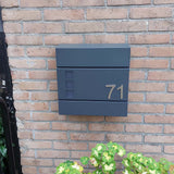 Le numéro Animo en acier inoxydable de 7,6 cm (sans perçage) de yessential.fr apparaît sous la forme « 71 » sur une boîte aux lettres moderne gris foncé, montée sur un mur de briques au-dessus de plantes vertes et jaunes.