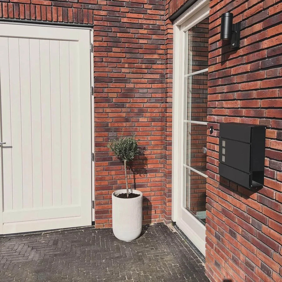 Une maison moderne en brique avec une porte blanche, une grande fenêtre, une boîte aux lettres Nevio Anthracite de yessential.fr, un petit arbre en pot dans une jardinière blanche et une lampe d'extérieur noire.