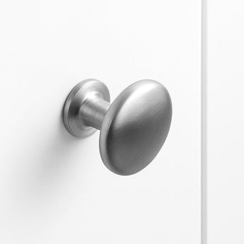 Le bouton de porte Pedro en acier inoxydable de yessential.fr présente un design ovale en métal brossé, monté sur une porte blanche unie et présenté en gros plan avec un style minimaliste et un éclairage doux.