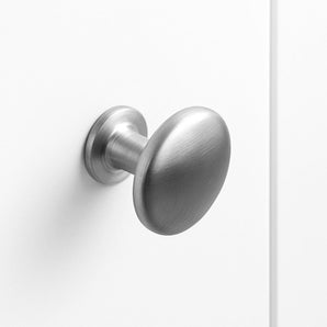 Le bouton de porte Pedro en acier inoxydable de yessential.fr présente un design ovale en métal brossé, monté sur une porte blanche unie et présenté en gros plan avec un style minimaliste et un éclairage doux.