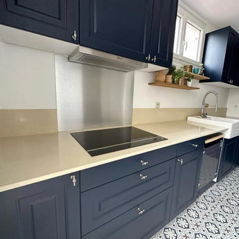 Cuisine moderne avec armoires bleu marine avec bouton Rando en acier inoxydable de yessential.fr, plan de travail beige, plaque de cuisson électrique, dosseret et acier inoxydable, évier de ferme, sol carrelé à motifs et étagères au-dessus avec plantes et pot et ustensiles.