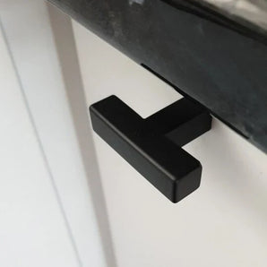 Gros plan du bouton noir Vigio de yessential.fr - une poignée moderne en forme de T d'un noir élégant - montée sur une porte d'armoire blanche sous un comptoir sombre.