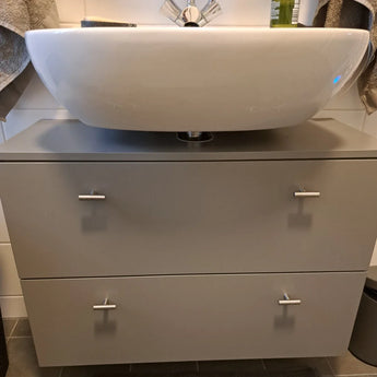 Gros plan d'un lavabo de salle de bain moderne sur un meuble sous-vasque gris avec deux tiroirs, chacun équipé du bouton Remo en acier inoxydable de yessential.fr pour un look élégant. Les serviettes sont visibles des deux côtés en arrière-plan.