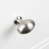 Le bouton Agero en acier inoxydable Deluxe de yessential.fr présente une finition en nickel brossé, un dessus rond légèrement aplati et une tige courte, présenté monté sur une surface blanche d'armoire ou de tiroir.