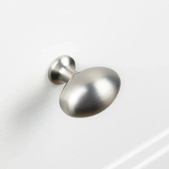 Le bouton Agero en acier inoxydable Deluxe de yessential.fr présente une finition en nickel brossé, un dessus rond légèrement aplati et une tige courte, présenté monté sur une surface blanche d'armoire ou de tiroir.