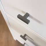 Gros plan d'une commode blanche avec des poignées Vigio Gunmetal Knob de yessential.fr, vue de dessus sous un angle. Les boutons modernes en forme de T accentuent la surface lisse et sont un exemple de design contemporain. Le sol apparaît en arrière-plan.