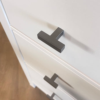 Gros plan d'une commode blanche avec des poignées Vigio Gunmetal Knob de yessential.fr, vue de dessus sous un angle. Les boutons modernes en forme de T accentuent la surface lisse et sont un exemple de design contemporain. Le sol apparaît en arrière-plan.