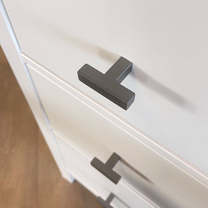 Gros plan d'une commode blanche avec des poignées Vigio Gunmetal Knob de yessential.fr, vue de dessus sous un angle. Les boutons modernes en forme de T accentuent la surface lisse et sont un exemple de design contemporain. Le sol apparaît en arrière-plan.