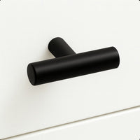Le bouton Remo Black de yessential.fr présente un design minimaliste en forme de T en noir mat, ajoutant une élégance moderne à une façade de tiroir blanche et lisse.