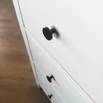 Gros plan d'un buffet blanc avec des boutons de meuble Marizo Black de yessential.fr en finition mate, présentés en angle sur un parquet, soulignant son design minimaliste.