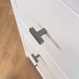 Gros plan de deux boutons Vigio Gunmetal de yessential.fr sur des tiroirs d'armoire blancs avec un plancher en bois en arrière-plan, pour une touche de style de bouton de meuble moderne.
