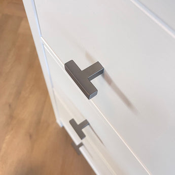 Gros plan de deux boutons Vigio Gunmetal de yessential.fr sur des tiroirs d'armoire blancs avec un plancher en bois en arrière-plan, pour une touche de style de bouton de meuble moderne.