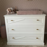 Une commode blanche à trois tiroirs avec une luxueuse finition dorée et des poignées yessential.fr Vasco Gold Knob est recouverte d'un matelas à langer rose, de peluches et d'articles de puériculture, contre un mur texturé et à motifs.