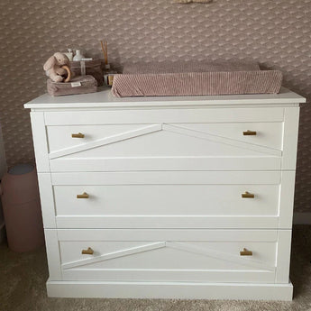 Une commode blanche à trois tiroirs avec une luxueuse finition dorée et des poignées yessential.fr Vasco Gold Knob est recouverte d'un matelas à langer rose, de peluches et d'articles de puériculture, contre un mur texturé et à motifs.