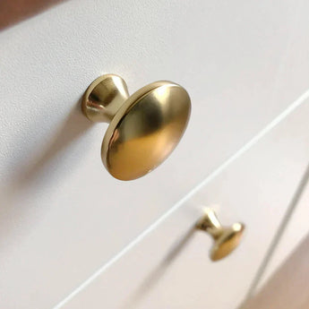 Gros plan de deux boutons d'armoire 10x Antoni Matte Gold de yessential.fr sur une armoire blanche, photographiés sous un angle pour montrer la finition lisse et brillante.