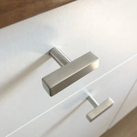 Gros plan d'un bouton moderne en acier inoxydable Vasco en métal brossé de yessential.fr, monté sur une façade de tiroir blanche avec un comptoir en bois visible sur le bord supérieur.